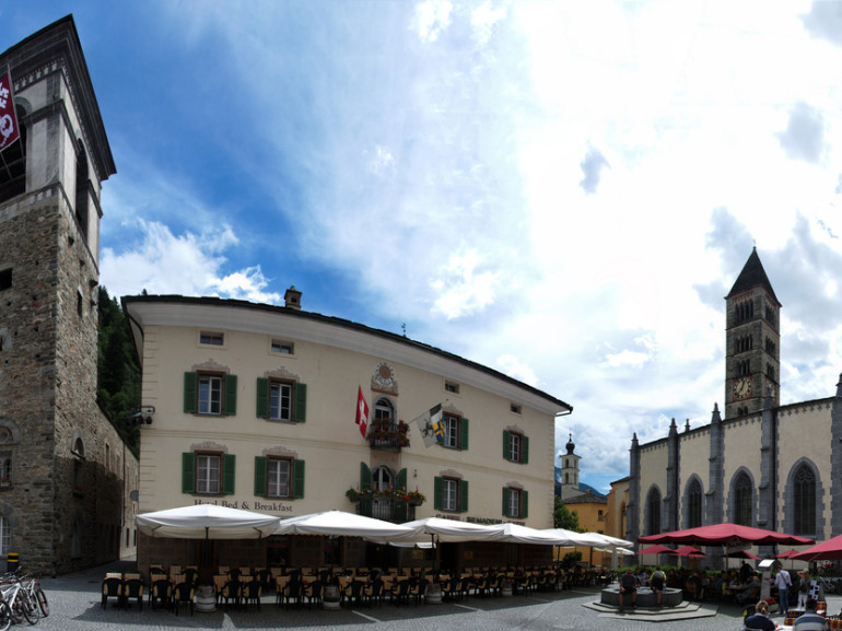 la piazza della città con il campanile in sasso e i tavolini del bar
