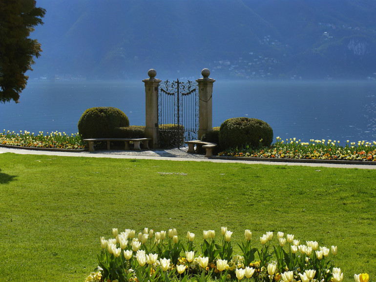 vista lago da un verde parco con aiuole fiorite. sul lago si apre un antico cancello in ferro battuto