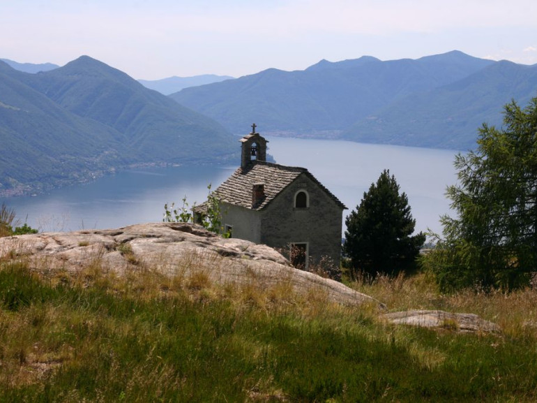 un chiesetta in mezzo ai prati con alle spalle il panorama sul lago maggiore