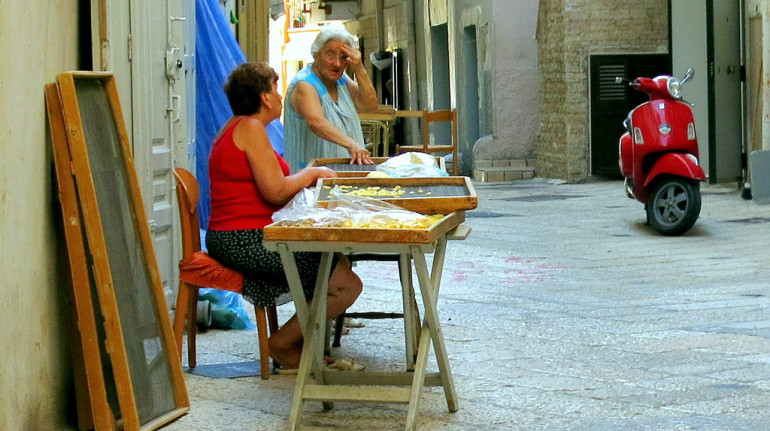 una via di bari con due signore sedute davanti a casa. su un banchetto vendono pasta fresca ai passanti